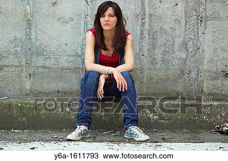 Stock Image - Portrait of a serious young woman looking at camera sitting in front of a cement wall. Fotosearch