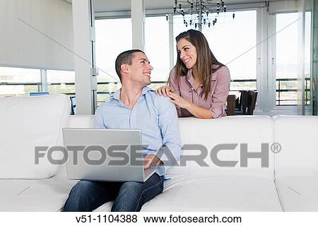 Stock Photo - Portrait of a young couple using laptop on the sofa in the living room. Fotosearch