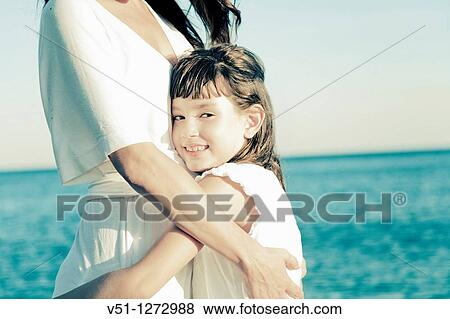 Stock Photo - Portrait of a young girl embracing her mother and looking at camera at the beach. Fotosearch