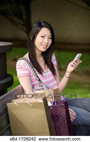 Portrait of a young woman sitting on a park bench with her cell phone and shopping bags View Large Photo Image Stock Image - Portrait of a young woman sitting on a park bench with her cell phone and shopping bags. Fotosearch