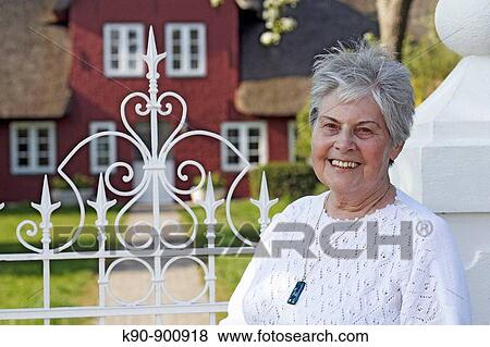 Stock Photo - Portrait of elderly caucasian woman at sunny weather smiling and looking into the camera with traditional Northfrisian house in the background, Island of Amrum, Schleswig_Holstein, Northern Germany, Europe. Fotosearch