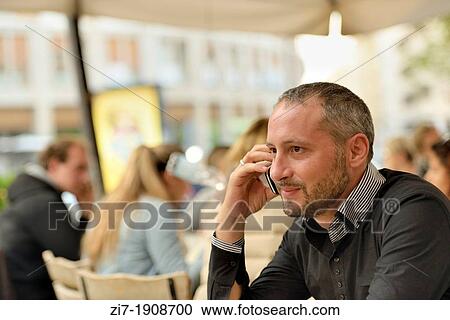Stock Image - portrait of man making a phone call with a cell phone, shot at happy hour time in a outdoor coffee bar, blurred in background, in city center. Fotosearch