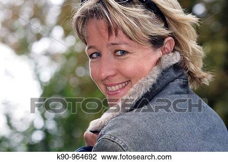 Stock Image - Portrait of young caucasian woman with sun glasses and short blond hair, smiling and looking into the camera, holding her hands at the collar, Hamburg, Germany, Europe. Fotosearch