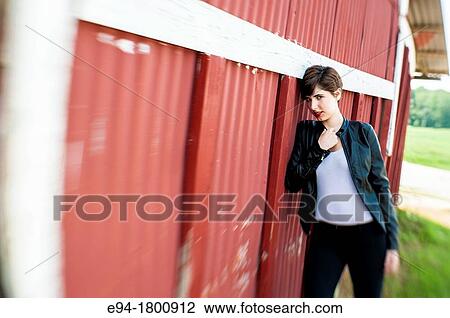 Stock Image - Portrait of young woman standing next to a red wall of a barn. Fotosearch