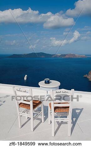 Postcard scene of two lonely chairs on terrace with table ready for tourists in Santorini Greece in Greek Islands View Large Photo Image Stock Photo - Postcard scene of two lonely chairs on terrace with table ready for tourists in Santorini Greece in Greek Islands. Fotosearch