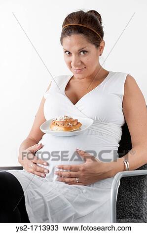Stock Image - Pregnant woman holding hands on her stomach and resting a plate with a sweet bun on her belly. Fotosearch