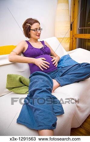 Stock Image - Pregnant woman sitting on a couch holding her belly. Fotosearch