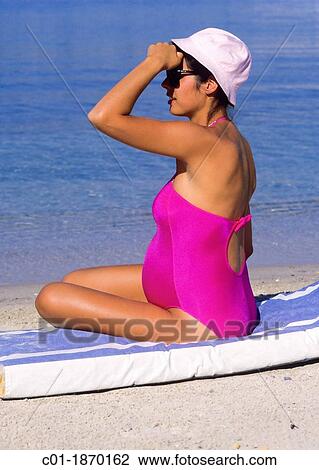 Stock Image - Pregnant woman sitting on beach and looking out to sea. Fotosearch