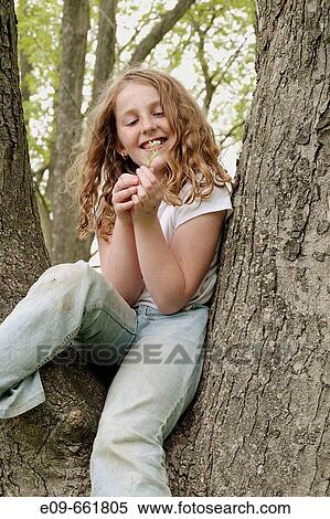 Stock Photography - Preteen girl with wavy gold-brown hair and freckles, sitting in tree, in faded jeans and white short-sleeved top, looking at small white wildflower and smiling. Fotosearch