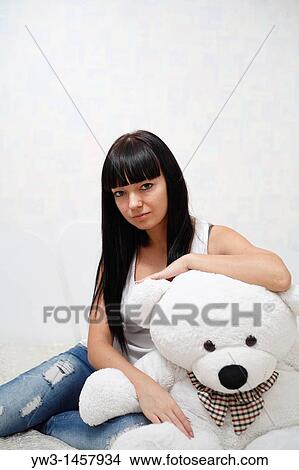 Picture - pretty brunette girl indoors embracing with big teddy bear on sofa. Fotosearch