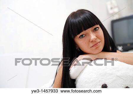 Stock Image - pretty brunette girl indoors embracing with big teddy bear on sofa. Fotosearch