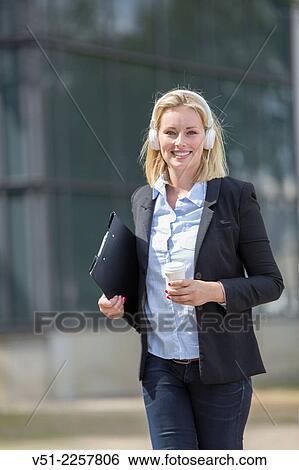 Stock Photograph - Pretty executive woman walking with coffee and headphones outdoors and smiling at camera. Fotosearch