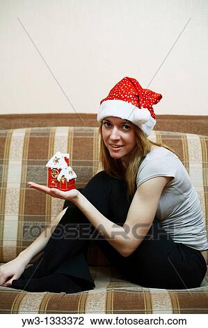 Stock Image - pretty girl in the Christmas hat on sofa with little red xmas house in hands. Fotosearch