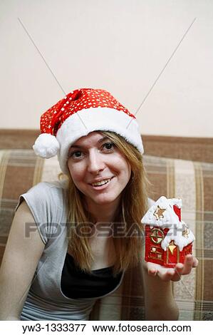 Stock Photo - pretty girl in the Christmas hat on sofa with little red xmas house in hands. Fotosearch