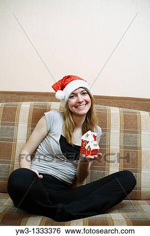 Stock Photograph - pretty girl in the Christmas hat on sofa with little red xmas house in hands. Fotosearch