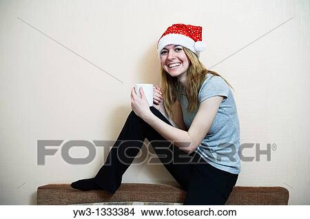 Picture - pretty girl in the Christmas hat against light wall with cup of tea or coffee. Fotosearch