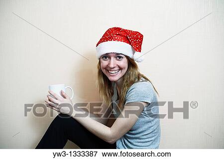 Stock Photo - pretty girl in the Christmas hat against light wall with cup of tea or coffee. Fotosearch