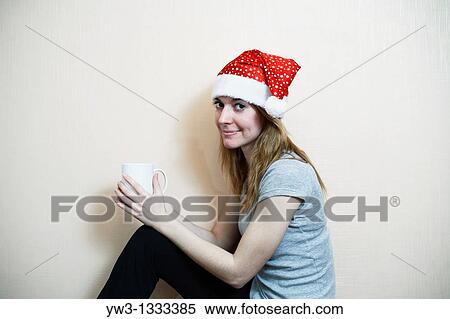 Stock Photography - pretty girl in the Christmas hat against light wall with cup of tea or coffee. Fotosearch