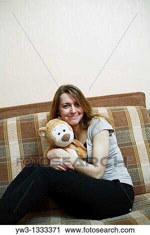 Stock Image - pretty girl sitting on sofa with teddy bear embracing and smiling. Fotosearch