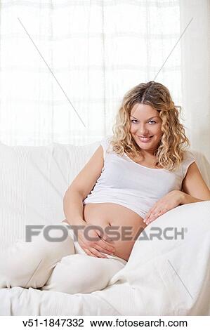 Stock Image - Pretty pregnant woman at sofa smiling at camera. Fotosearch