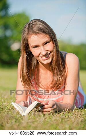 Stock Photo - Pretty young woman reading a book in park and smiling at camera. Fotosearch