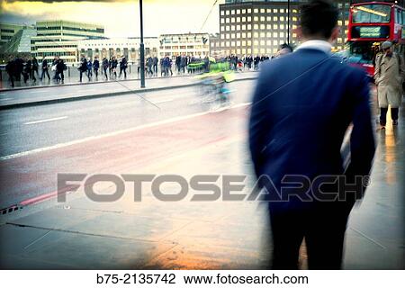 Stock Image - Rear view of a close up unrecognizable man walking along London Bridge and many people walking in the background. London, England, UK, Europe. Fotosearch