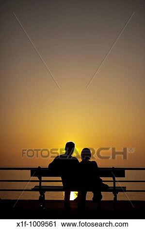 Rear view of a young romantic young couple sitting on a seaside bench watching the sun setting over the sea, Aberystwyth Wales UK View Large Photo Image Stock Image - Rear view of a young romantic young couple sitting on a seaside bench watching the sun setting over the sea, Aberystwyth Wales UK. Fotosearch