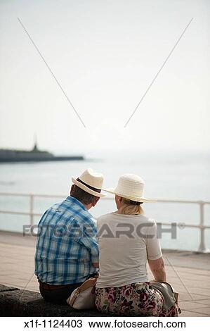 Stock Image - Rear view of an adult senior couple, man woman, sitting outside wearing hats, Summer afternoon, Aberystwyth Ceredigion wales UK. Fotosearch