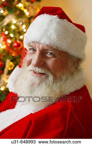 Stock Image - Santa looks at the camera in front of a Christmas tree. Fotosearch
