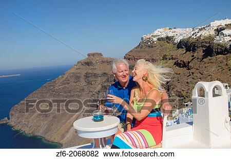 Stock Image - Santorini Greece retired couple on holiday having fun having wine on cliff in Fira Greek vacation.. Fotosearch