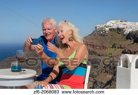 Stock Image - Santorini Greece retired couple on holiday having fun with cell phone pictures on cliff in Fira Greek vacation.. Fotosearch