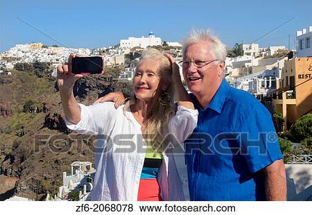 Stock Photo - Santorini Greece retired couple on holiday having fun with cell phone pictures on cliff in Fira Greek vacation.. Fotosearch