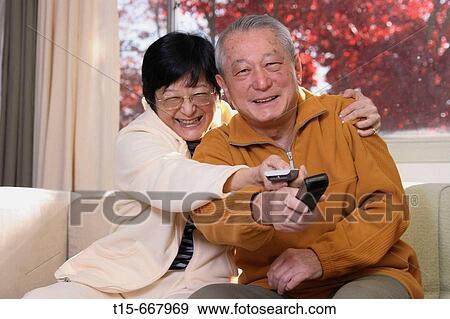 Stock Photo - Senior Asian couple having some silly fun to watch their favorite program on tv using the remotes. Fotosearch