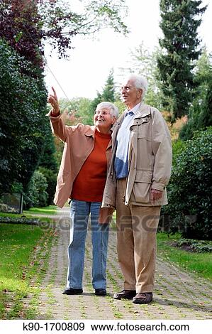 Stock Photo - Senior caucasian couple standing besides each other, looking up into the air while the woman is pointing with her finger at something, blurred green background, Hamburg, Germany, Europe. Fotosearch