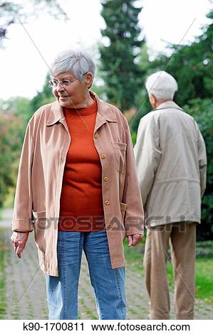 Stock Image - Senior caucasian woman, wearing glasses, looking down with senior man behind her, seen from backside, blurred green background, Hamburg, Germany, Europe. Fotosearch