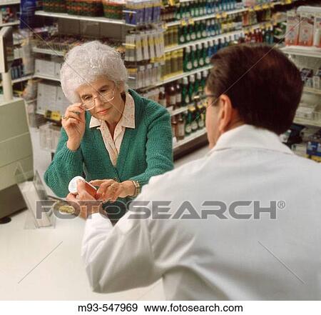 Stock Photo - Senior woman adjusting glasses and reading the label on a prescription drug bottle, which is being handed to her by a pharmacist. Pharmacy shelves with product visible in the background. Fotosearch