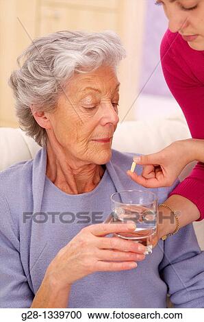 Stock Image - senior woman in a mauve cardigan, holding a glass of water, and young help giving her a whited tablet at home. Fotosearch