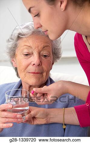 Senior woman in a mauve cardigan, holding a glass of water, and young help giving her a red tablet at home View Large Photo Image Stock Image - Senior woman in a mauve cardigan, holding a glass of water, and young help giving her a red tablet at home. Fotosearch