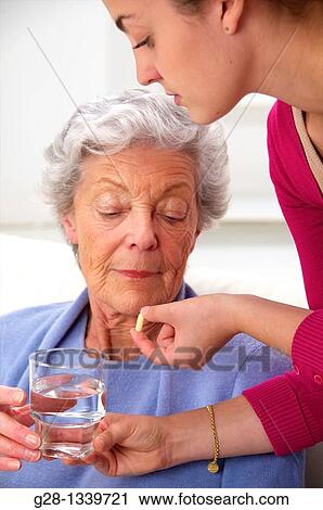senior woman in a mauve cardigan, holding a glass of water, and young help giving her a white tablet at home View Large Photo Image Stock Image - senior woman in a mauve cardigan, holding a glass of water, and young help giving her a white tablet at home. Fotosearch
