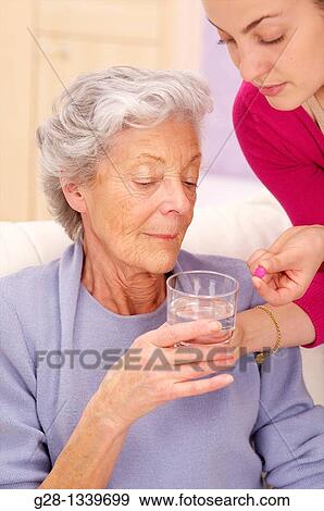 senior woman in a mauve cardigan, holding a glass of water, and young help giving her a red tablet at home View Large Photo Image Stock Photo - senior woman in a mauve cardigan, holding a glass of water, and young help giving her a red tablet at home. Fotosearch