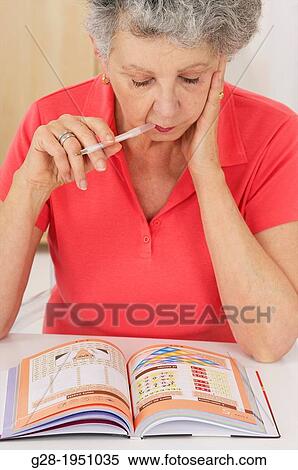 Stock Photography - Senior woman in red thinking over a test memory book for Alzheimer with a pen in her mouth. Fotosearch