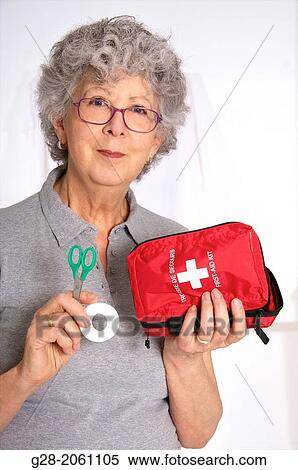 Senior woman, or senior nurse, with red glasses, in grey, holding green scissors and roll of plasters in one hand and red first aid kit in the other, at home or in surgery View Large Photo Image Stock Photography - Senior woman, or senior nurse, with red glasses, in grey, holding green scissors and roll of plasters in one hand and red first aid kit in the other, at home or in surgery. Fotosearch