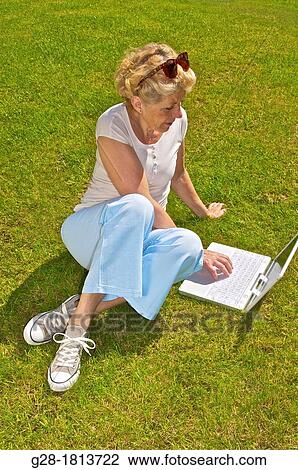 Stock Image - Senior woman sitting on the grass, and writing on her laptop. Fotosearch