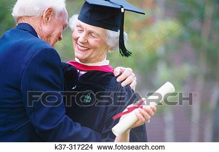 Picture - Senior woman wearing graduation cap and gown being congratulated by her husband on receiving degree. Fotosearch