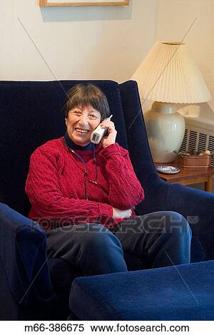 Stock Photography - Seventy-four year old woman sitting in a living room chair talking on a portable phone in her New York home.. Fotosearch