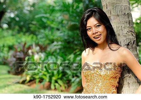 Stock Image - Sexy young woman, flirting, leaning agains tree trunk, Indonesia, Asia. Fotosearch