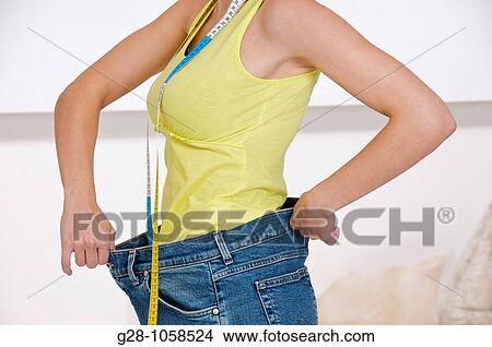 shoulder to hips portrait of a young woman holding her too large jeans with measuring tape around her neck View Large Photo Image Picture - shoulder to hips portrait of a young woman holding her too large jeans with measuring tape around her neck. Fotosearch