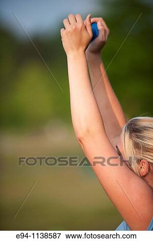Stock Photo - Side aview of a woman in workout clothes holding a dumbbell above her head at a boot camp.. Fotosearch