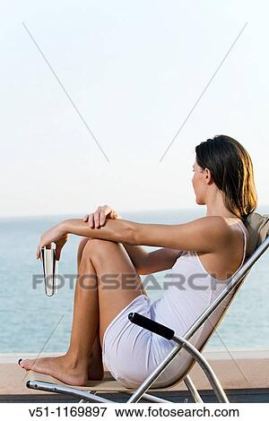 Stock Photo - Side view of a woman looking at the ocean from a terrace with a book. Fotosearch