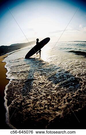 Stock Photograph - Silhouette of a surfer entering the water at sunrise. Fotosearch
